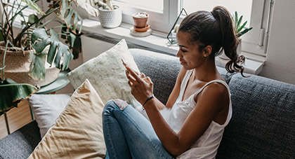 A young woman using her smartphone while sitting in a blue comfy couch to fill out eVisit questionnaire on the MyChart app.