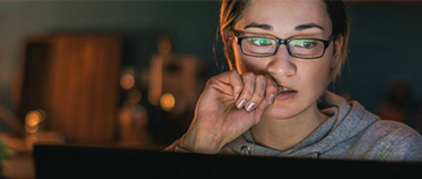 A woman with glasses, using her laptop in the evening to look at 24/7 Virtual Urgent Care telehealth video appointments.