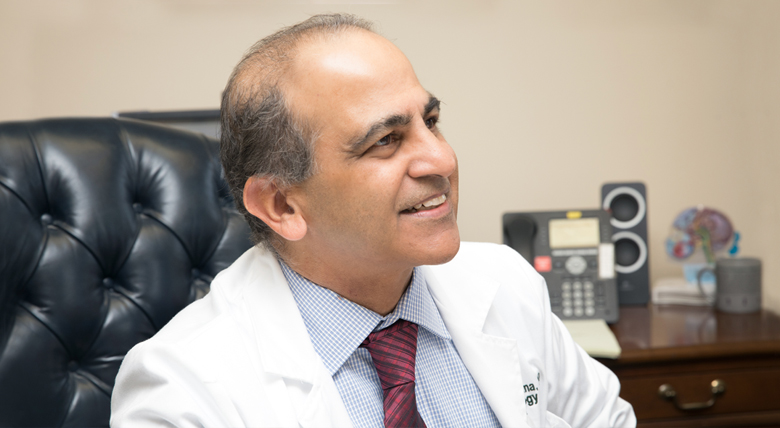 A medical professional, a South Asian man in a white coat, smiles while sitting at his desk.