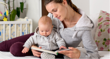 A mother on a bed and holding her baby while reading a book.