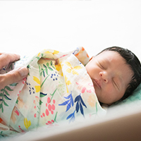 A newborn wrapped in a blanket sleeping in a bassinet with a parent's hand on the baby.