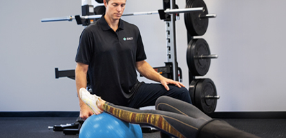 A medical professional helps a patient stretch on a blue exercise ball in a gym.