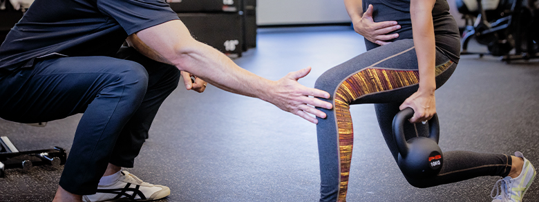 A medical professional in a black shirt helps a pregnant person do lunges with a kettlebell.
