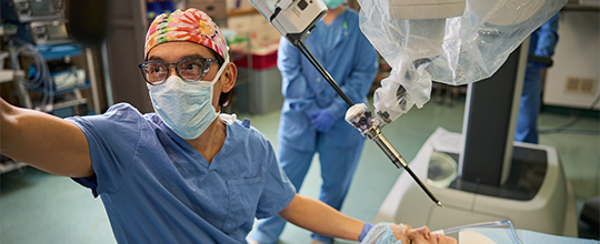 A medical professional in a blue scrub top and colorful cap is using a robotic arm during a surgery.