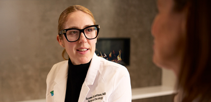 A female medical professional in a white coat and glasses speaks to a patient.