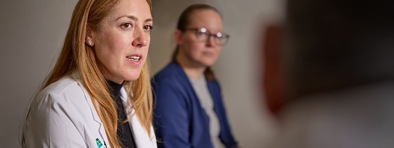 A female medical professional with reddish blonde hair in a white coat speaks intently in a meeting.
