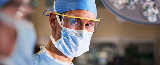 A medical professional in a surgical mask, blue scrubs, and safety glasses looks at the viewer.