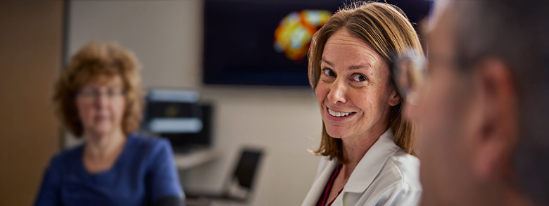 A female, medical professional with brown hair and a white coat smiles, looking toward the viewer.