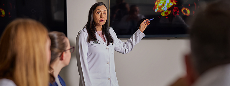 A female doctor in a white lab coat with the Allegheny Health Network (AHN) logo is actively presenting in a well-lit room.