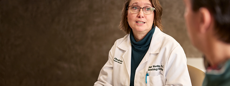 A female medical professional in glasses and a lab coat speaks earnestly to a person off-screen.