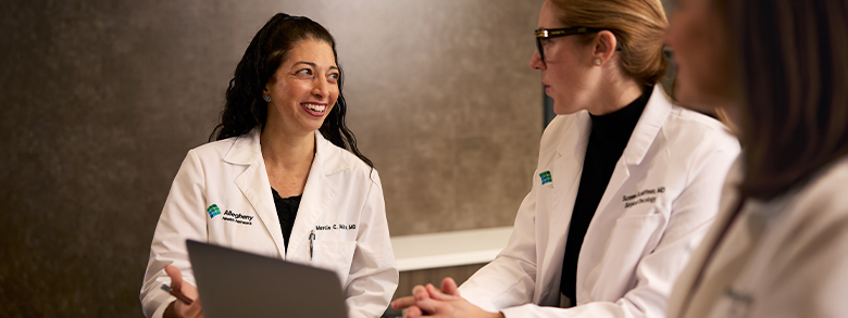Three female, medical professionals in white lab coats discuss something, one smiling brightly.