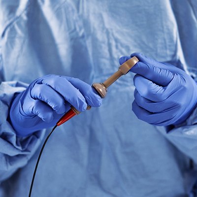 A female medical professional in blue scrubs looks up, holding a camera-like device in a medical setting.