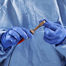 A female medical professional in blue scrubs looks up, holding a camera-like device in a medical setting.