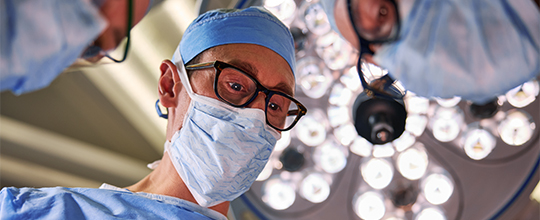 A medical professional in a blue cap, mask, and scrubs looks down intently during surgery.
