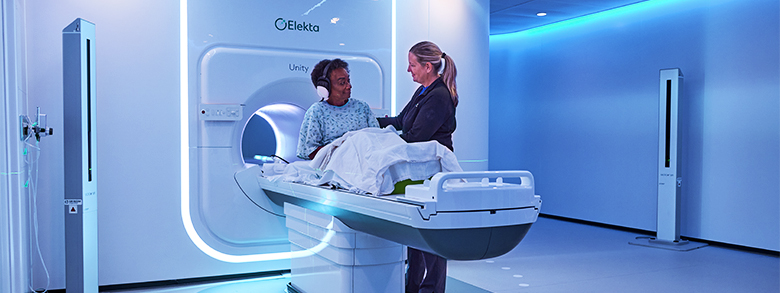 A medical professional in blue scrubs comforts a patient before they enter a large medical scanner.