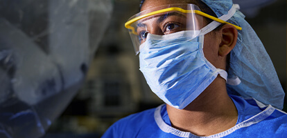 A female surgeon in blue scrubs, a surgical cap, mask, and glasses, stands in an operating room with medical equipment in the background.