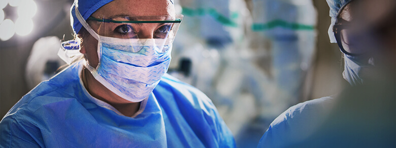 A female surgeon in blue scrubs, surgical cap, face mask, and eye protection looks intently forward in an operating room.