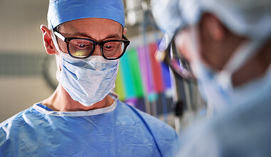 A male surgeon in blue scrubs, a surgical cap, face mask, and glasses looks intently down during an operation.
