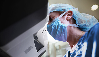 A female surgeon in blue scrubs, surgical cap, and face mask looks into a robotic surgical console.