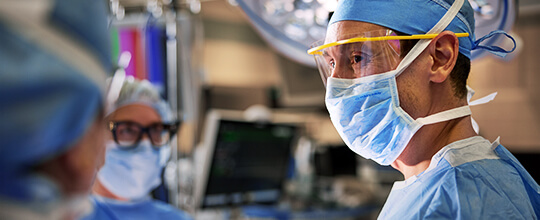 A surgeon in a blue scrub cap, face mask, and eye shield looks intently during an operation, with other medical staff visible in the foreground.