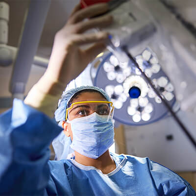 A female healthcare professional in a scrub cap, face mask, and eye protection adjusts an overhead surgical light.