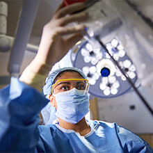 A female healthcare professional in a scrub cap, face mask, and eye protection adjusts an overhead surgical light.