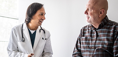 An Asian medical professional in a white coat smiles at an older white patient in a plaid shirt.