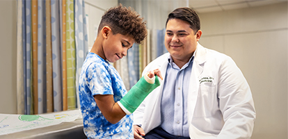 A young boy in a tie-dye shirt with a green arm cast talks to a medical professional in a white lab coat.