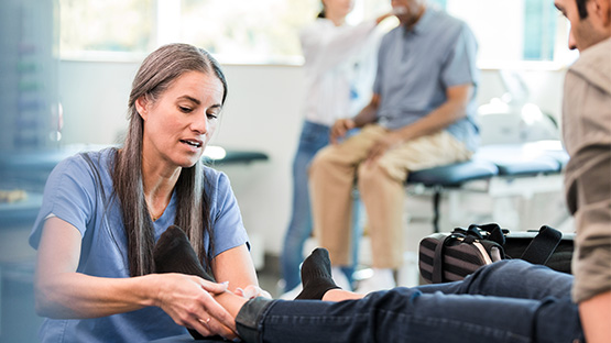 A physical therapist examines a patient's ankle while another patient receives care in the background.