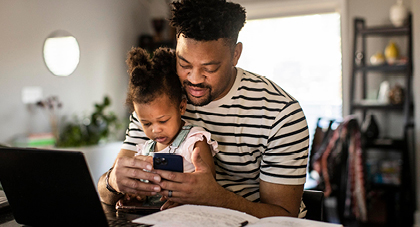 A father witting with his young daughter on his lap while checking an app on his phone.