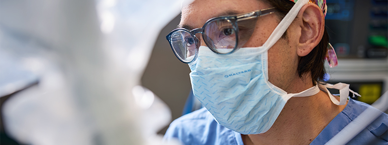 A medical professional in blue scrubs, a mask, and glasses looks focused during work.